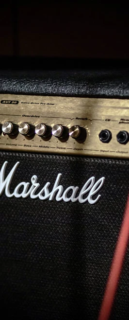 A close-up of a Marshall amplifier with red audio cables plugged in, glowing indicator light visible. A close-up of a Marshall amplifier with red audio cables plugged in, glowing indicator light visible.